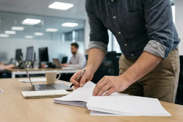 A person stands at a desk in a modern office, reviewing papers and holding a pen. A laptop, mug, and notebook are